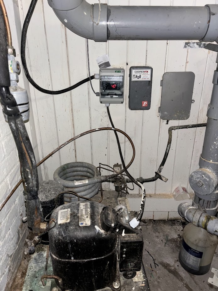 Commercial refrigeration compressor beside wall-mounted temperature controls, wiring and piping