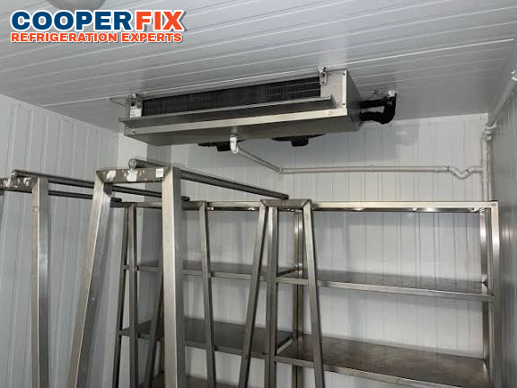 Ceiling-mounted evaporator above metal storage shelving in a cold room.